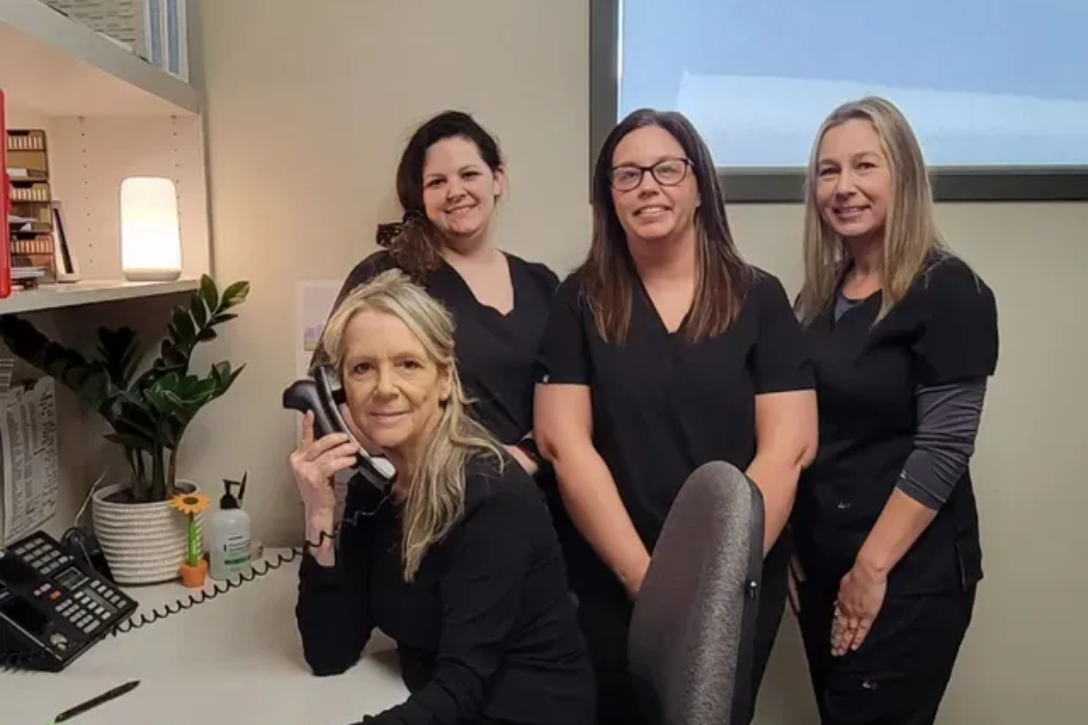 Four women in black scrubs behind a desk, one on the phone. Office setting.