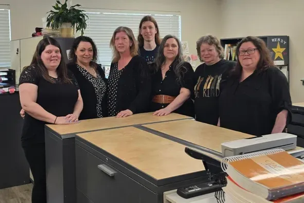 Seven women standing behind a desk in an office. They are wearing black clothing and smiling.