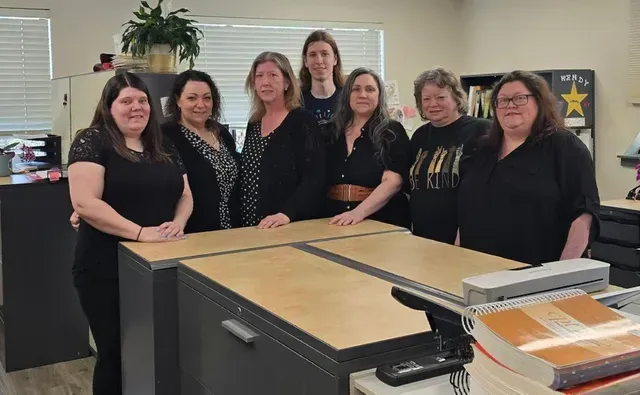 Seven people standing behind a desk in an office, most wearing black.