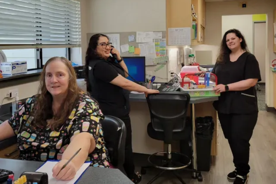 Three medical office staff members. One on phone, two at desks. Brightly lit room with supplies.