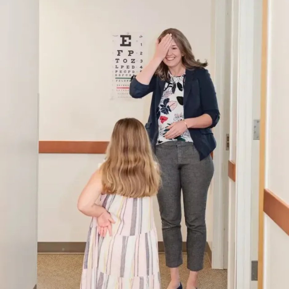 Woman testing a child's vision using an eye chart. The woman covers one eye, child stands in front.