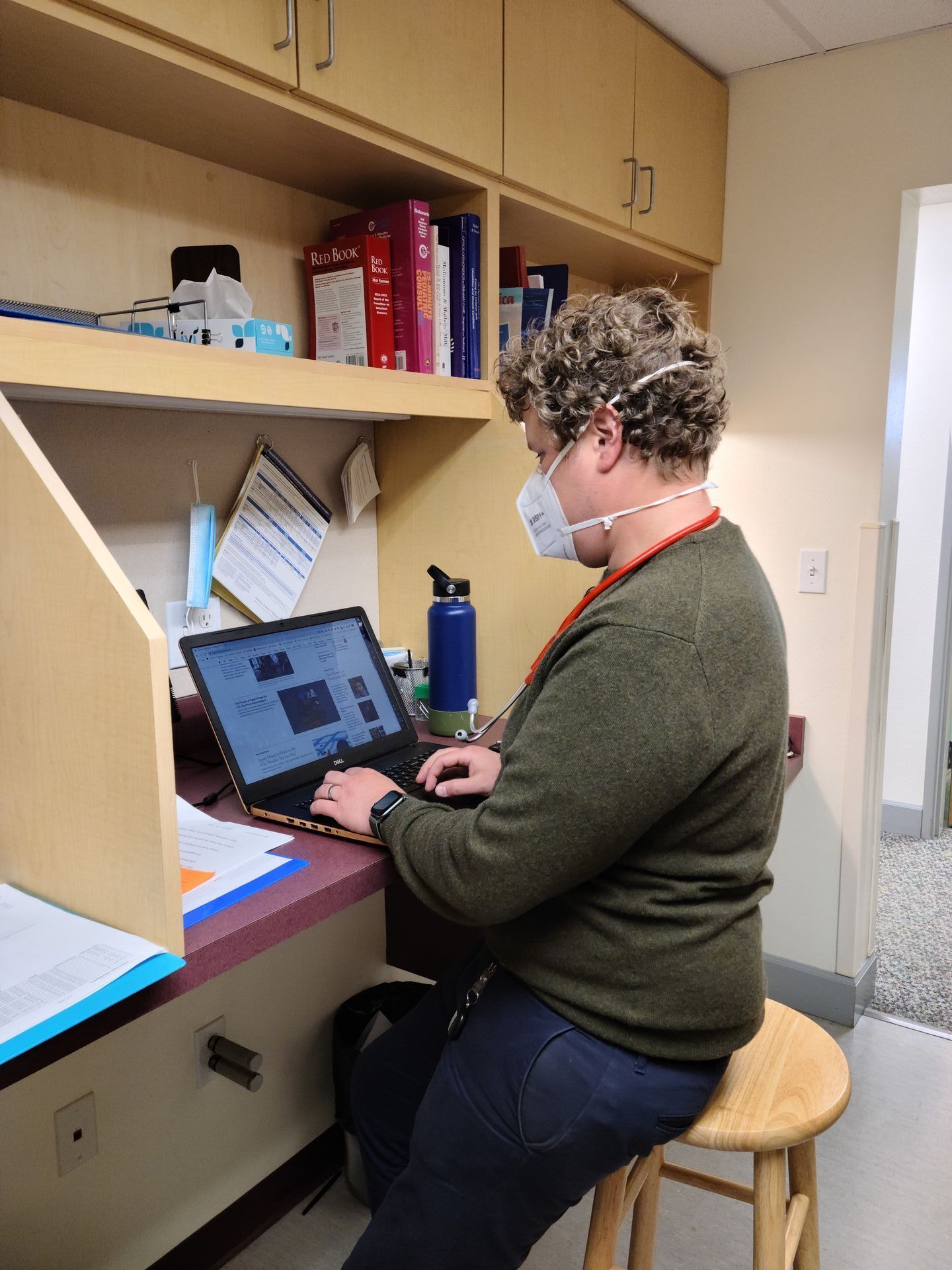 Person wearing a mask seated at a desk, typing on a laptop. A water bottle is behind the laptop.