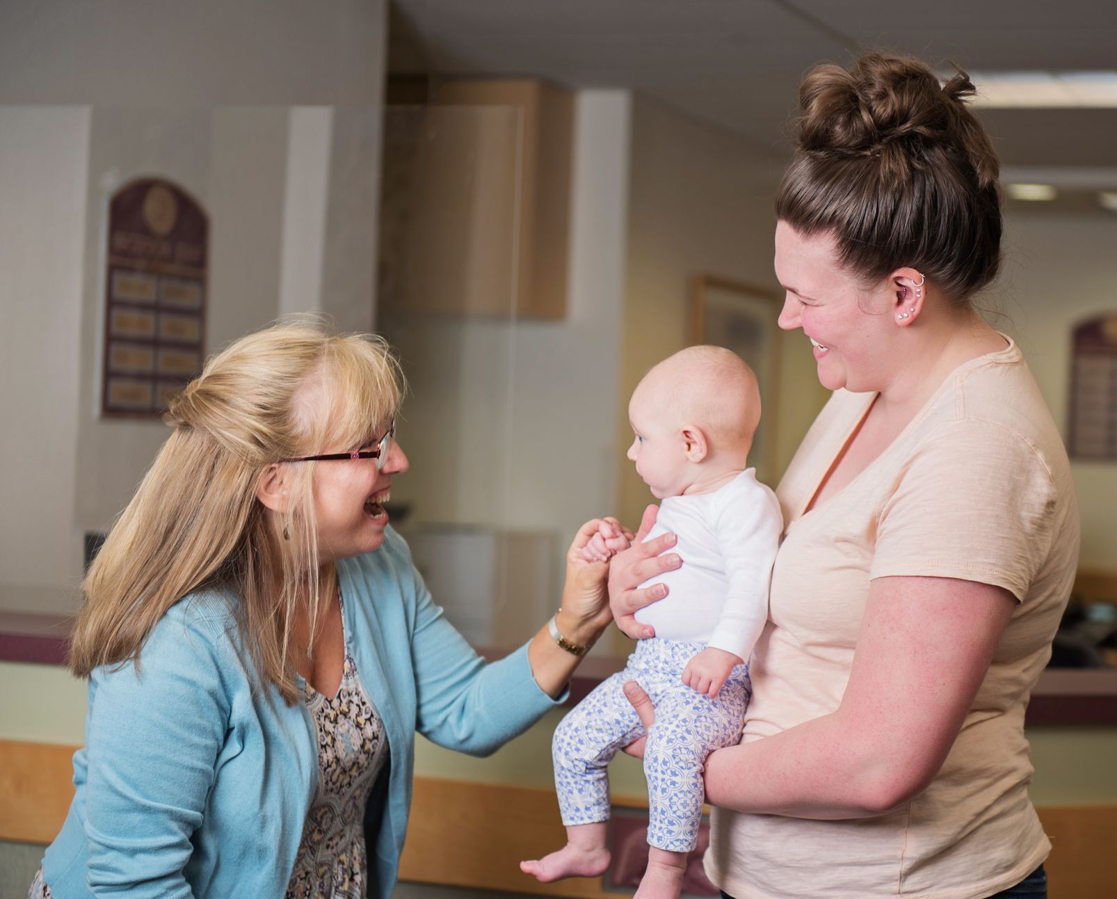 Woman holding baby, smiling at another woman in a hallway. The other woman smiles and touches the baby.