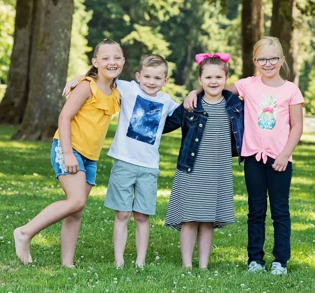 Four children smiling and hugging in a park. Green grass, trees, and bright clothing.