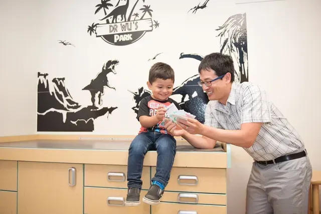A doctor smiles and interacts with a young child sitting on a medical table in an office with dinosaur decor.