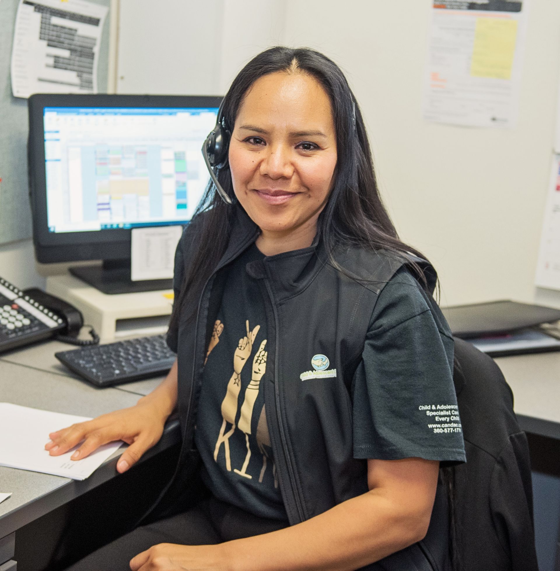 Woman wearing a black vest smiles at the camera, sitting at a desk with a computer and phone.