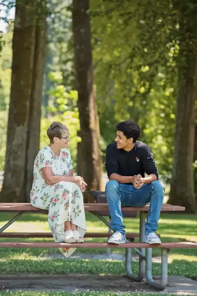 Woman and young person sitting on a park picnic table, talking. Outdoors, trees in background.