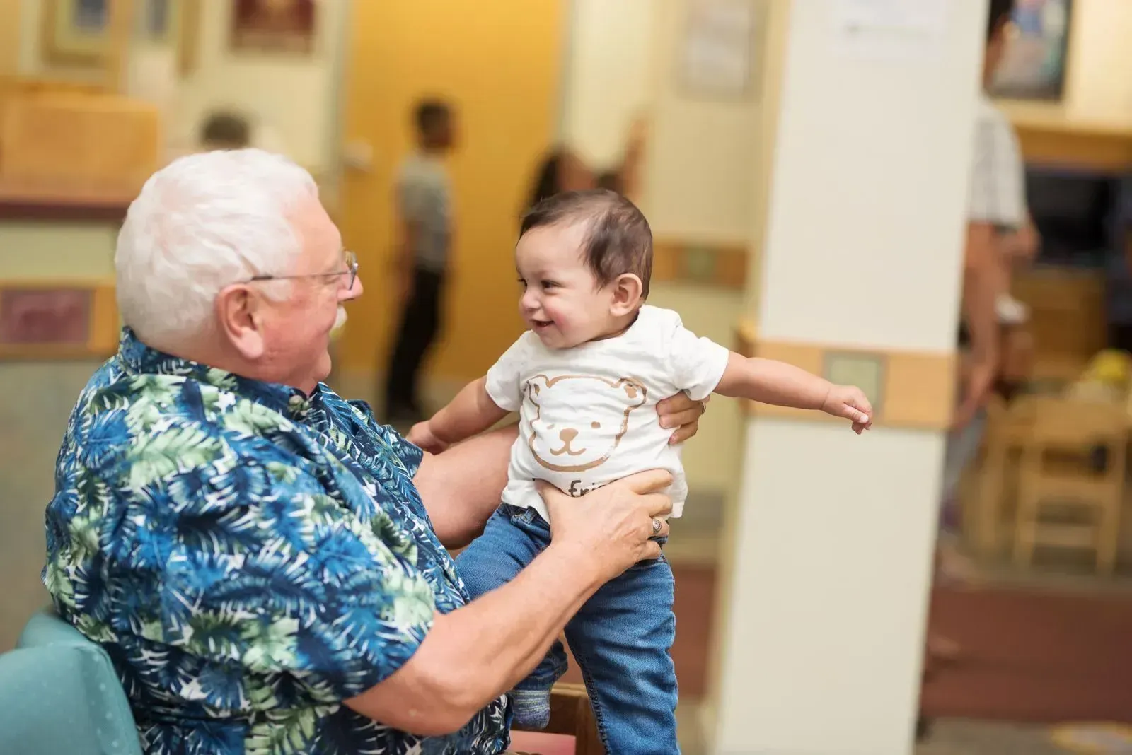 An older person holds up a smiling baby, arms outstretched, in a light-filled room.