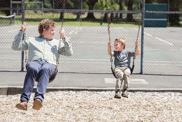 Man and child swinging on swings at a playground, gray fence and court in background.