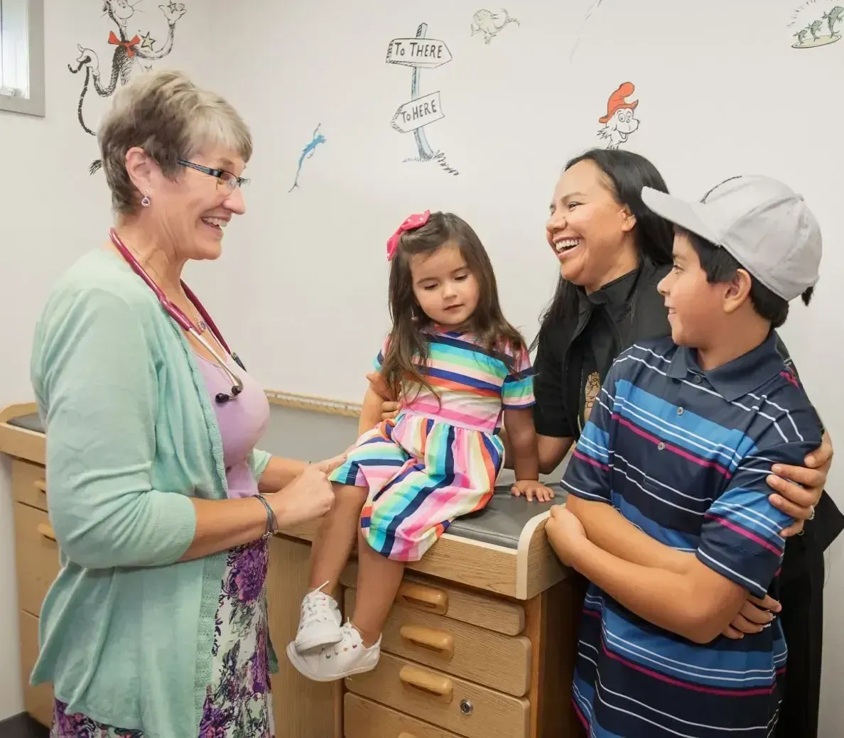 Doctor with stethoscope smiling at family in a clinic room; child sits on exam table.