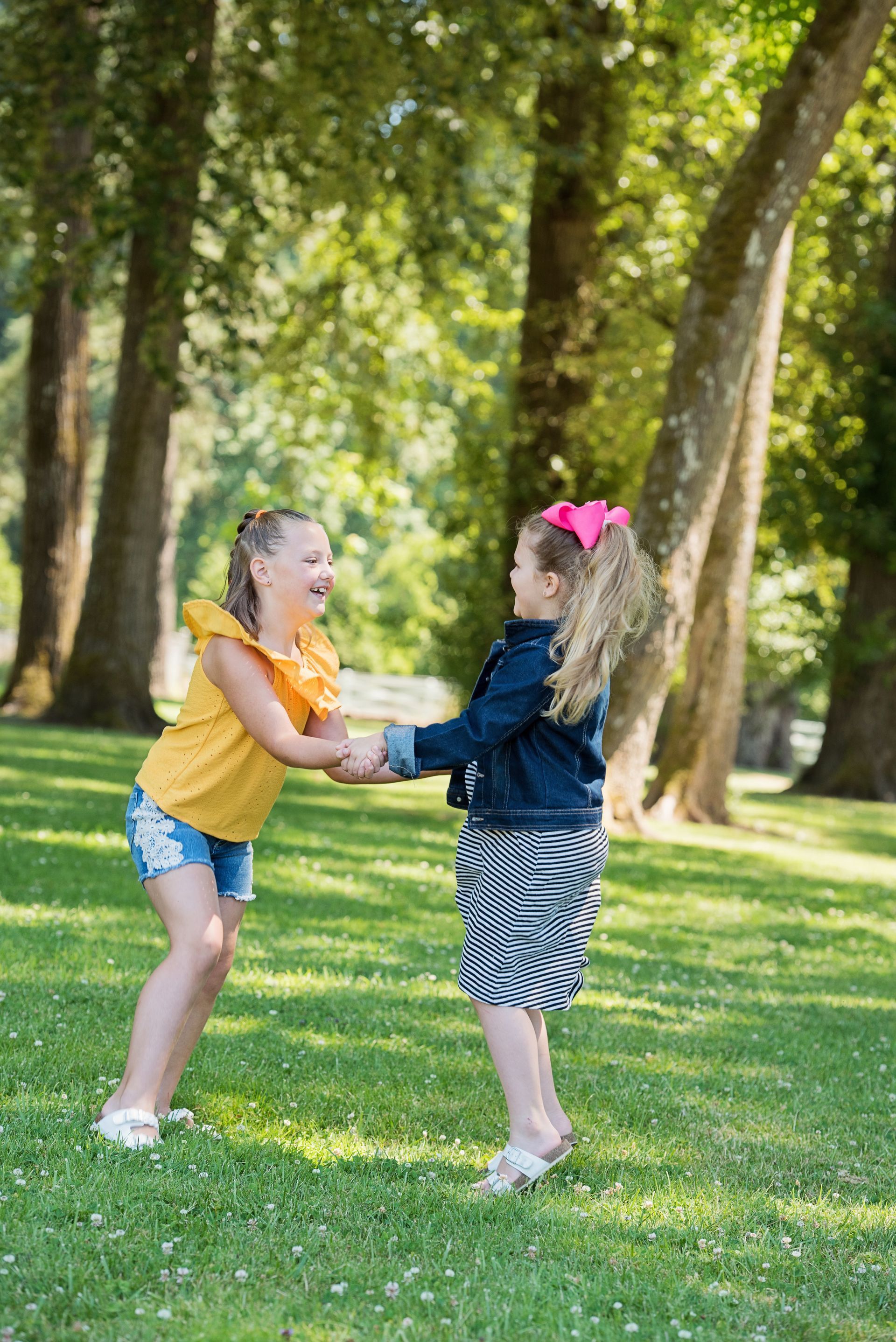 Two girls holding hands, laughing, in a grassy park, trees in background, sunny day.
