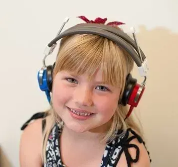 Girl with blonde hair wearing colorful hearing test headphones, smiling.