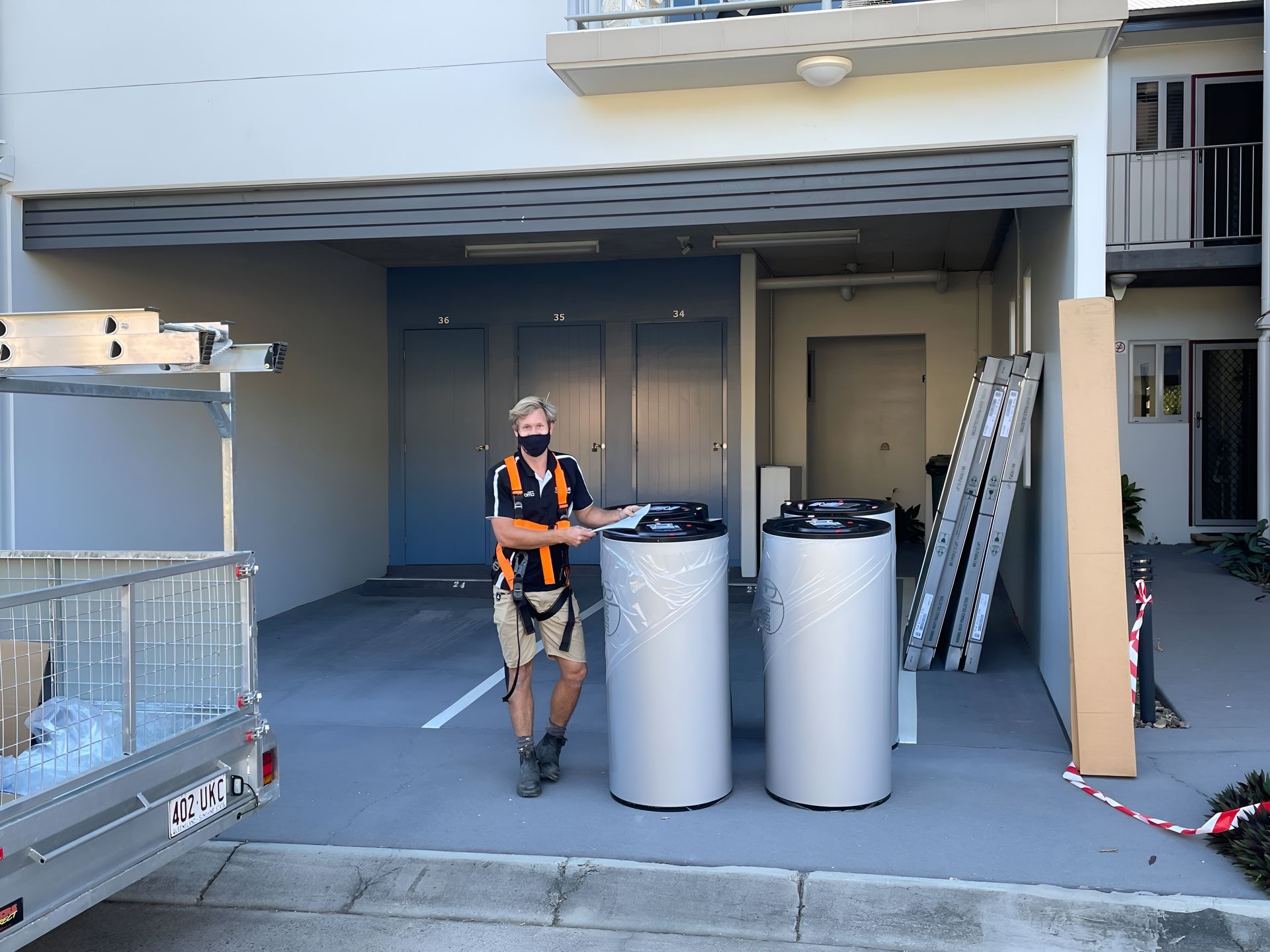 Man in Safety Harness Stands Beside Two Silver Water Heater — Taps 'R' Us in Maroochydore, QLD