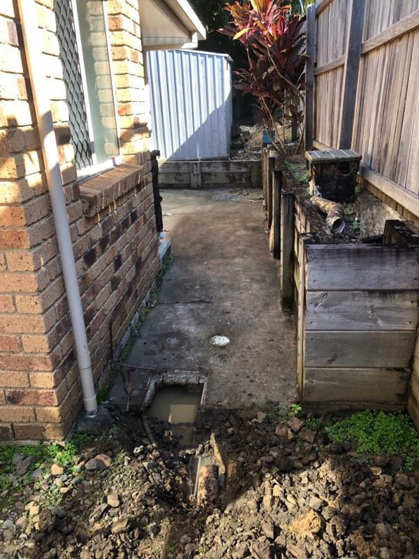 Narrow outdoor space with exposed concrete, bricks, wood fence, and dirt. A hole of water and soil.— Taps 'R' Us in Kunda Park, QLD