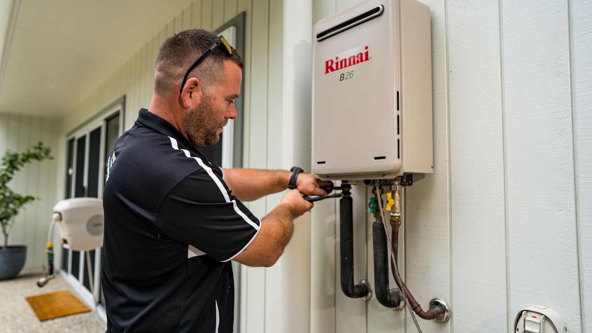 Man in a black shirt fixing a Rinnai B26 water heater on a house exterior. — Taps 'R' Us in Kunda Park, QLD