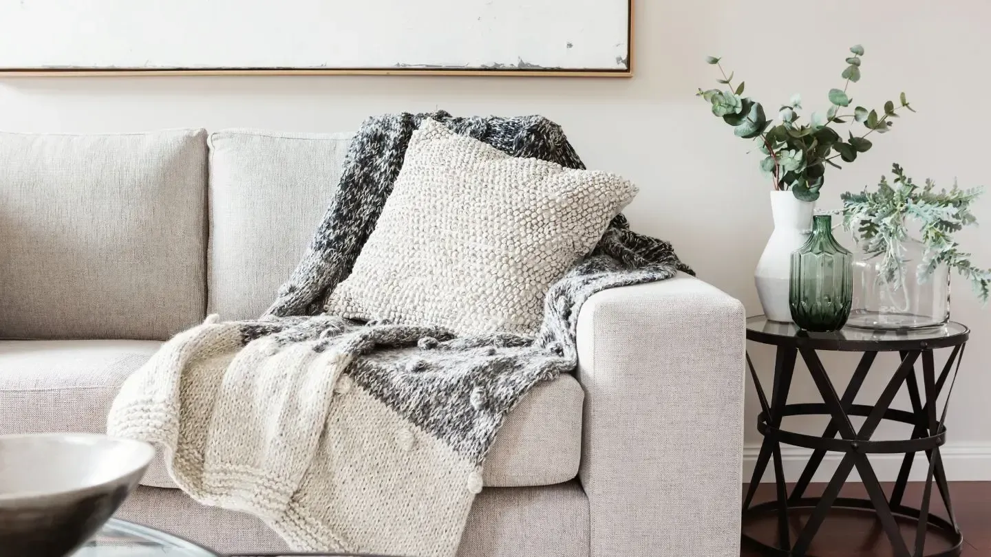 Cozy living room scene. Beige sofa with gray blanket, textured pillow. Side table with vase of greenery.