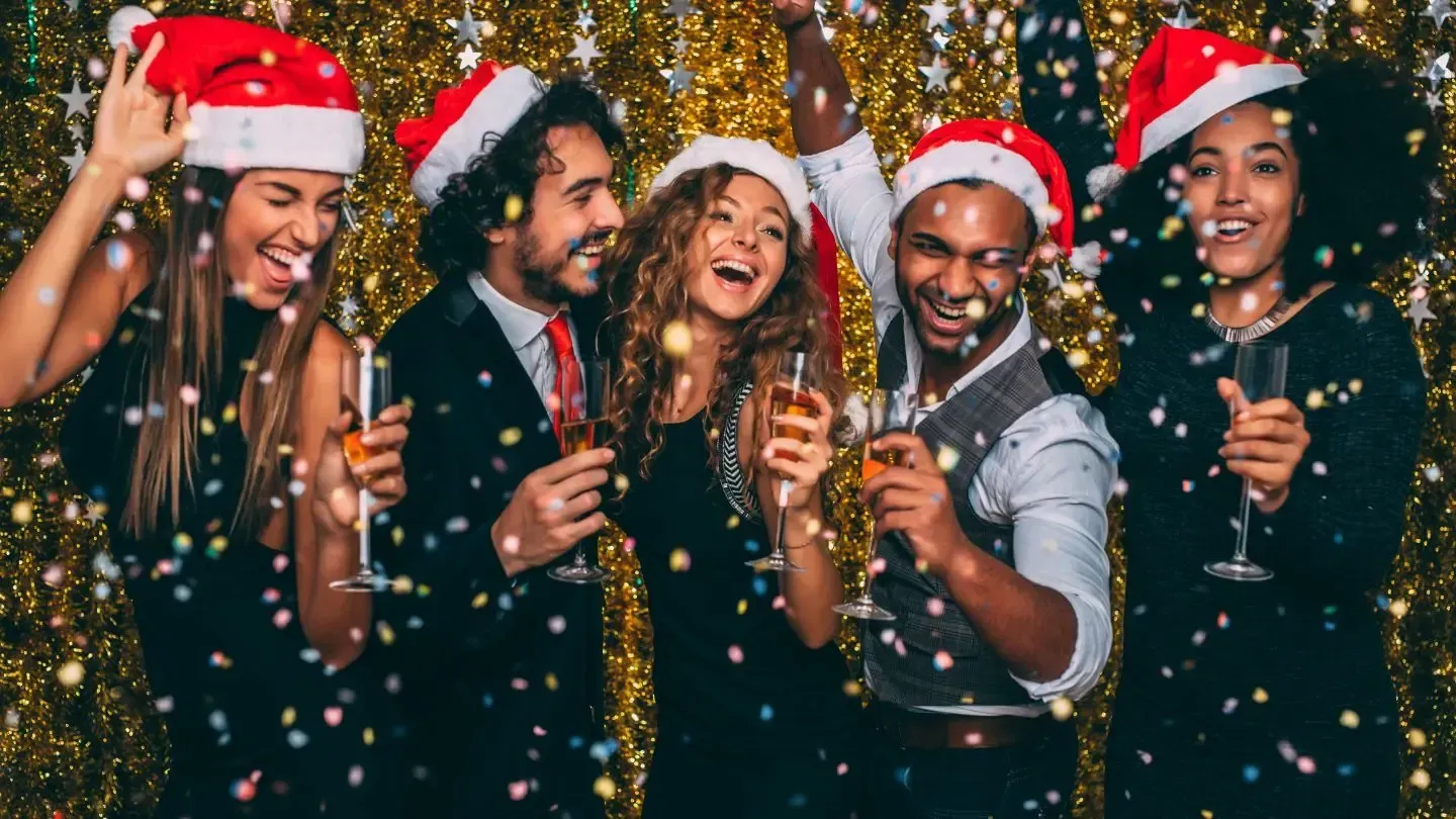 People celebrating with champagne and Santa hats, gold confetti, festive backdrop.