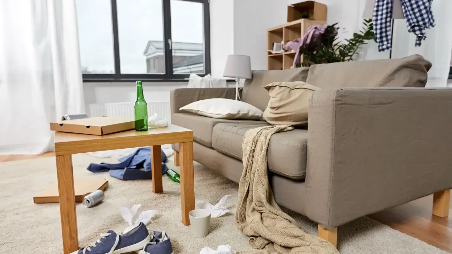 Messy living room with couch, coffee table, pizza box, beer bottle, and scattered items.