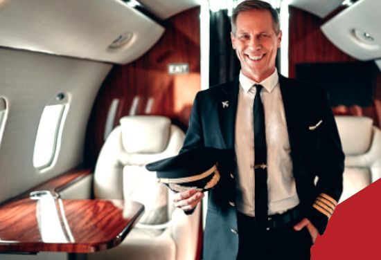 Smiling airline pilot standing in a private jet cabin, holding his cap and wearing a dark uniform.