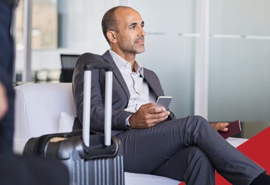 Man in a suit sitting on a couch, holding a phone beside a suitcase in an airport lounge.