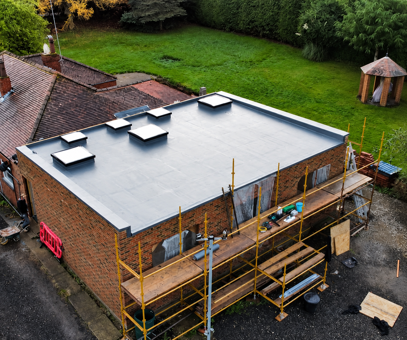 Rooftop renovation on a brick house with scaffolding, white roofing membrane, and green yard nearby