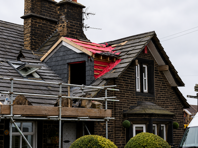 House with damaged red roof and smoke-stained chimney, showing storm damage under repair