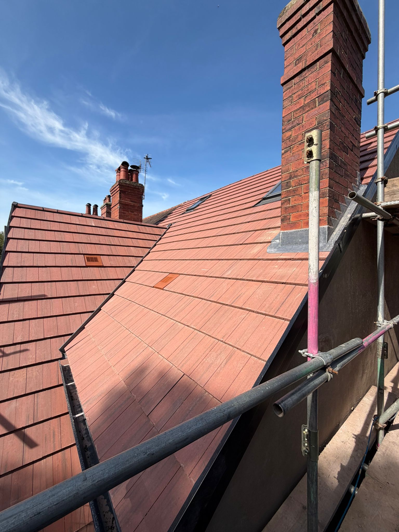 Red tiled rooftops and brick chimneys under a blue sky, seen from scaffolding during roof repairs