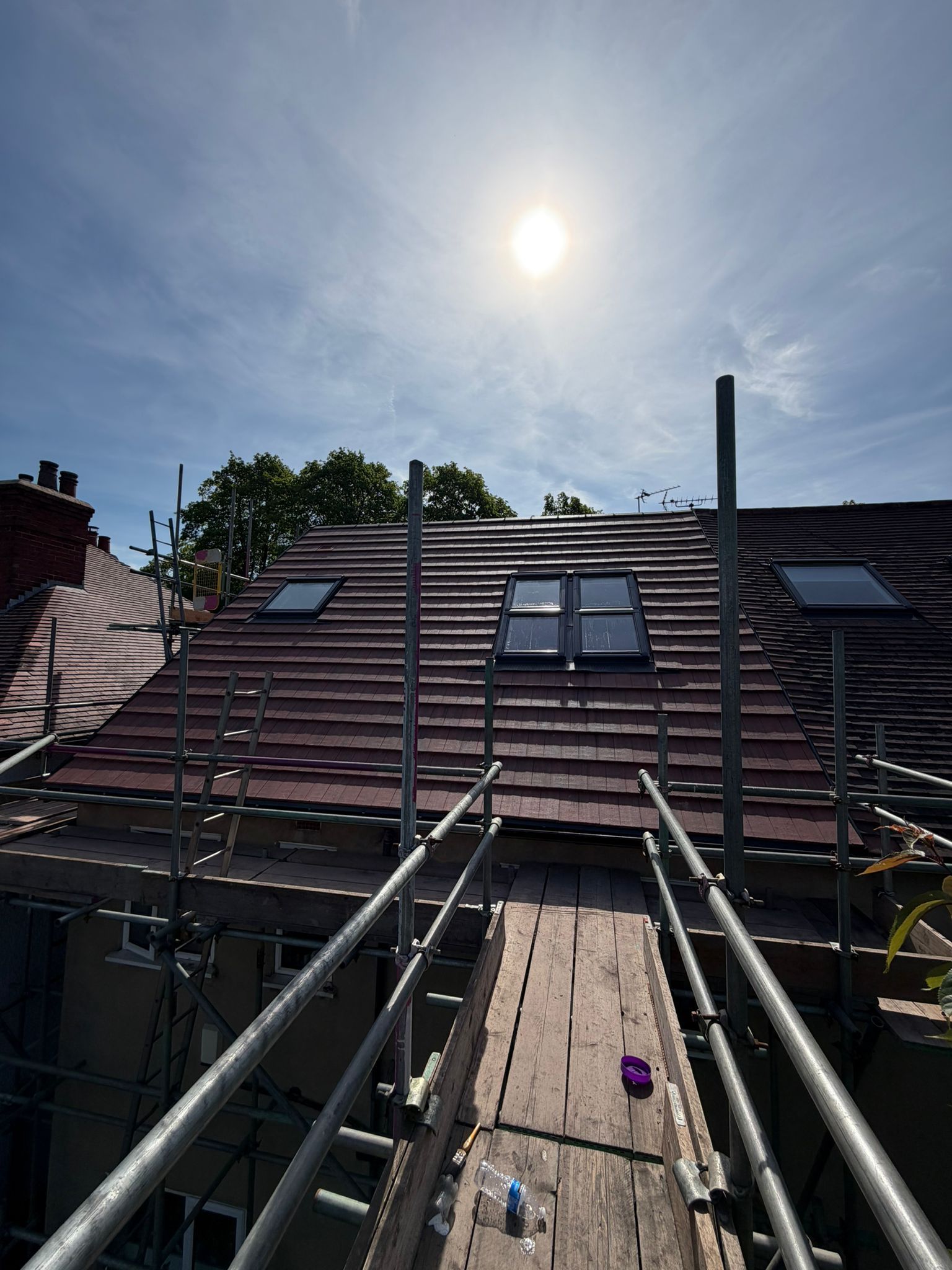 Scaffolding leading to a red-tiled roof under a bright sun, with solar panels and a cloudy blue sky
