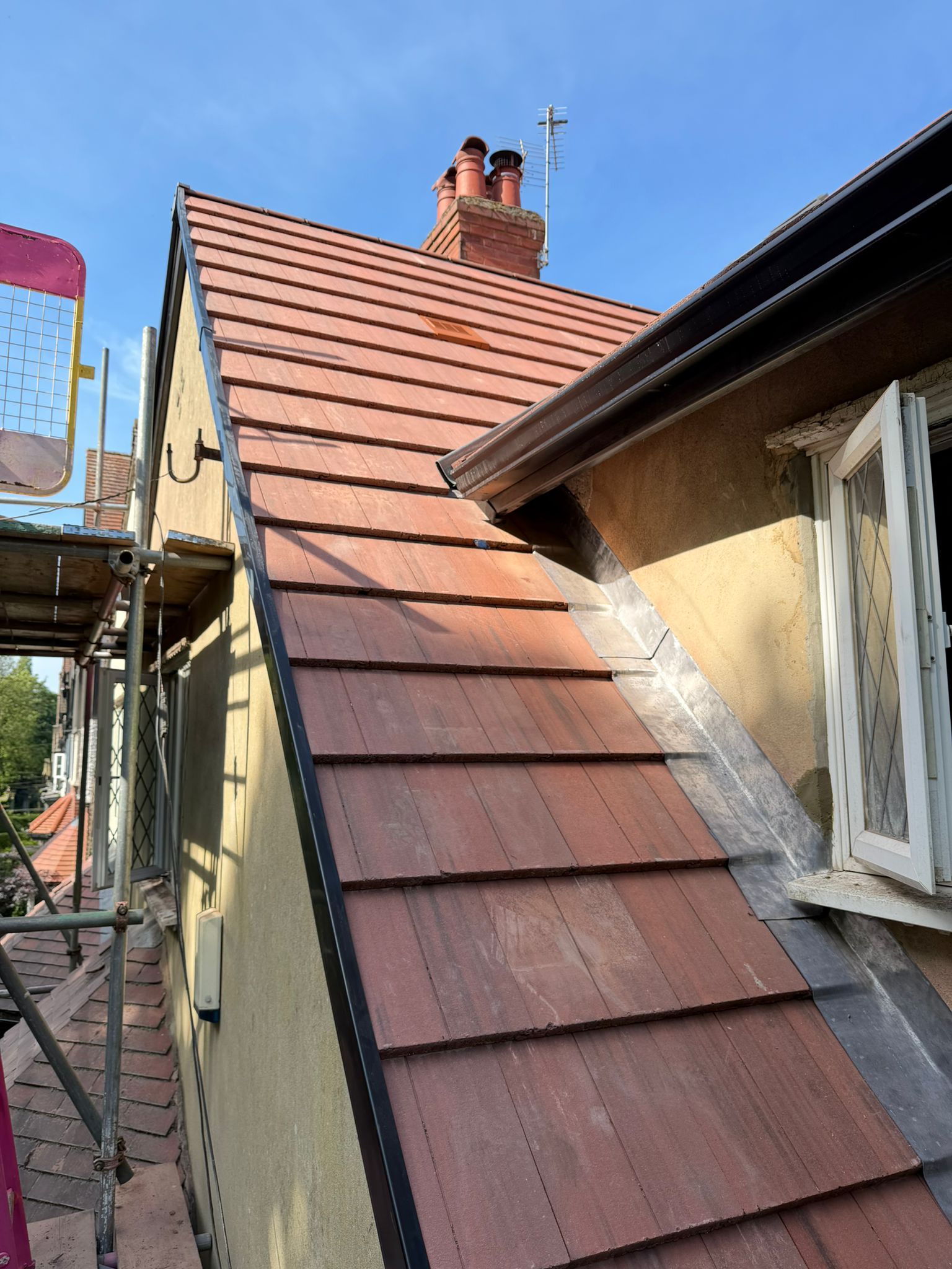Narrow house roof with red tiles and a chimney beside a white window under a clear blue sky