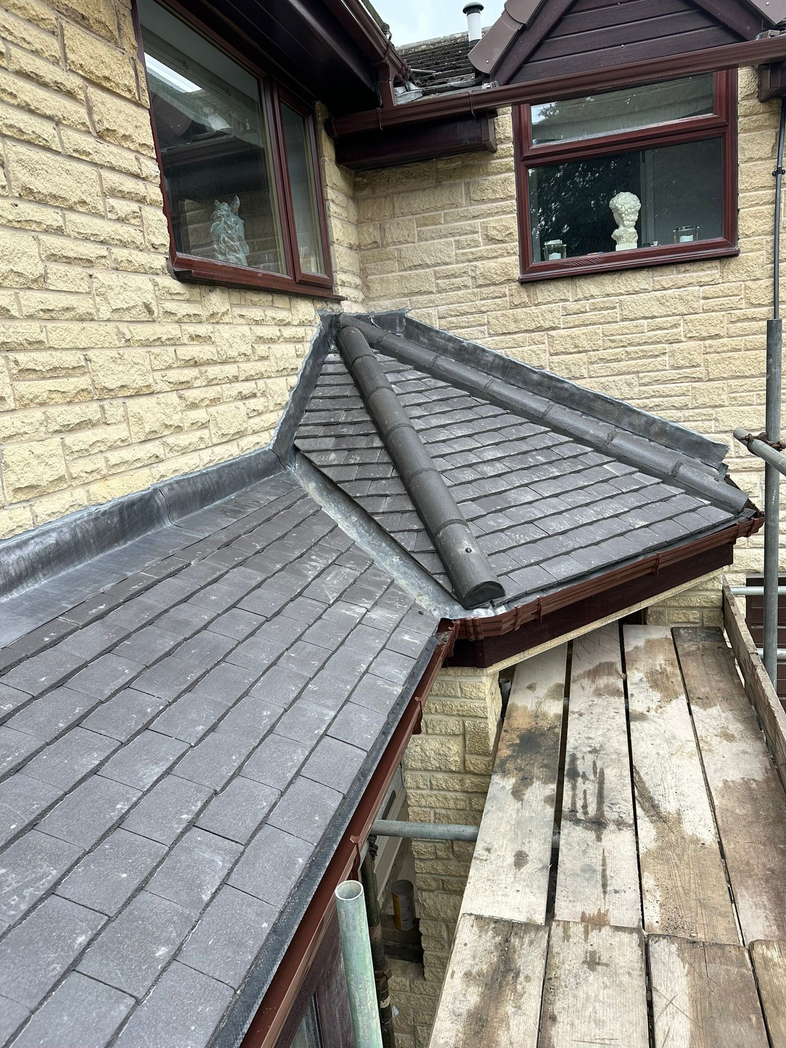 Roof repair with gray shingles and a wooden walkway beside a beige stone house