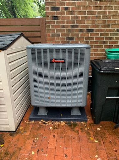 An air conditioner is sitting on a brick patio next to a shed and trash cans.