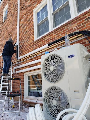 A man is standing on a ladder next to a large air conditioner on the side of a brick building.