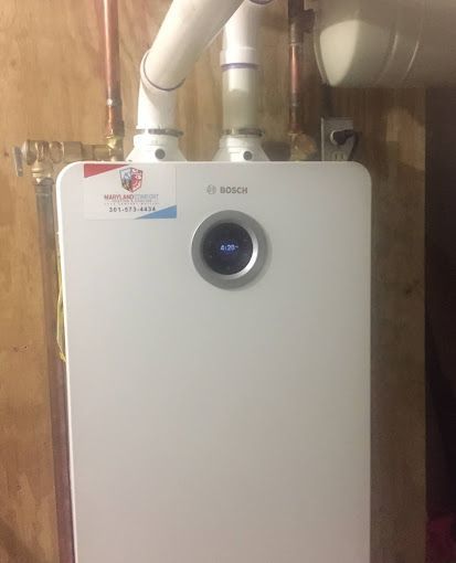 A white boiler is sitting on top of a wooden wall next to pipes.