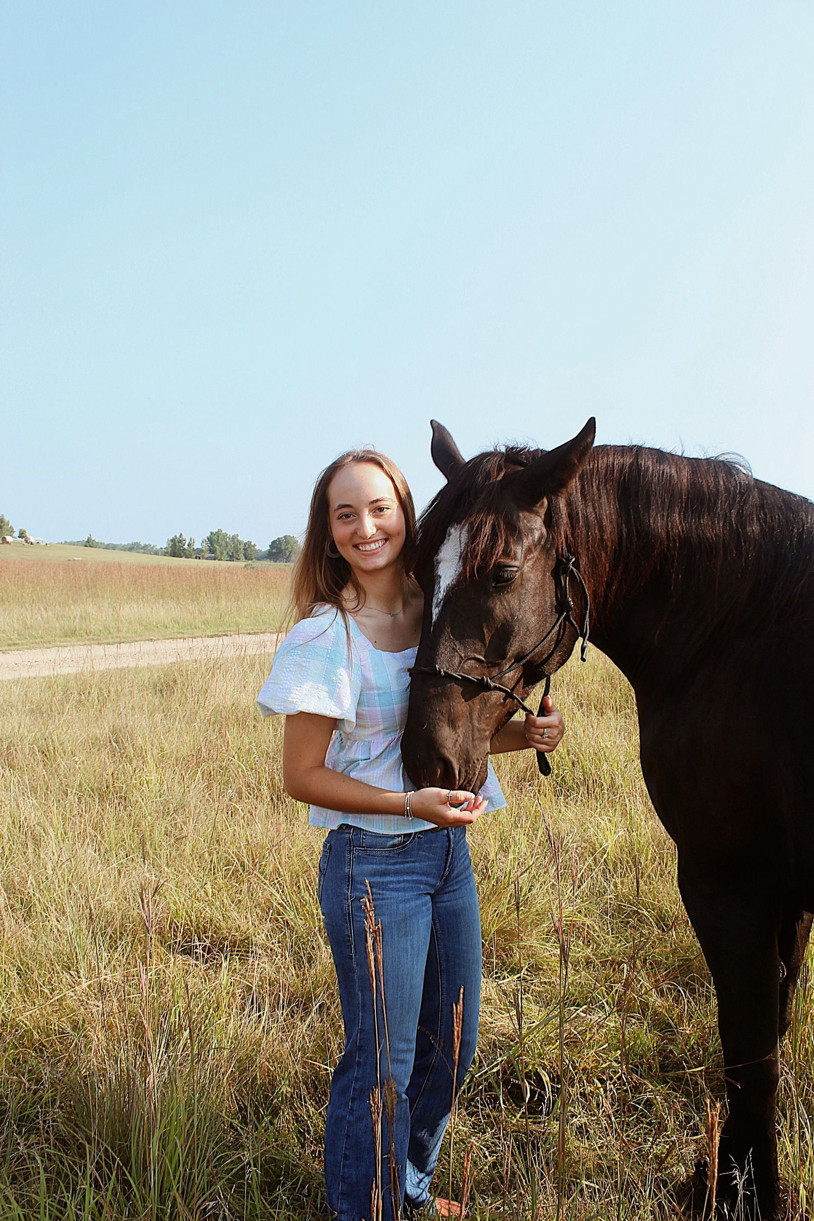 Woman in field with dark horse; blue top, jeans; sunny day.