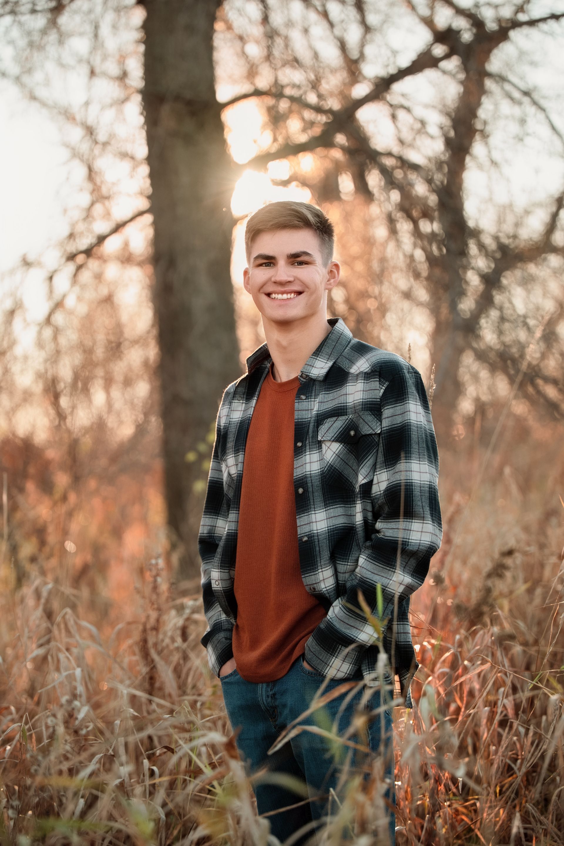 Young man in plaid shirt smiles outdoors at sunset, hands in pockets.