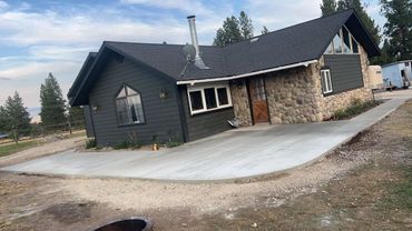 A one-story house with dark gray siding and a stone facade, featuring a newly poured concrete patio in a rural setting.