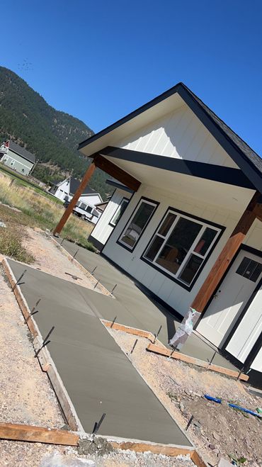 A newly poured concrete walkway leads to the entrance of a modern white house with dark trim, set against a mountain backdrop.