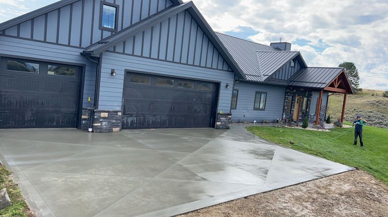 A newly poured concrete driveway leads to a large, blue-sided house with metal roofing, still under construction.