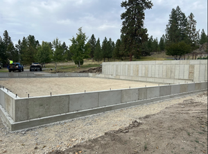 A concrete foundation for a new building sits on a gravel lot under a cloudy sky with trees in the background.