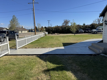 A new concrete walkway leads to a set of stairs at the entrance of a house, beside a white picket fence on a sunny day.