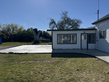 A white house with a flat-roofed extension, a newly poured concrete patio, and a pickup truck parked in the backyard.