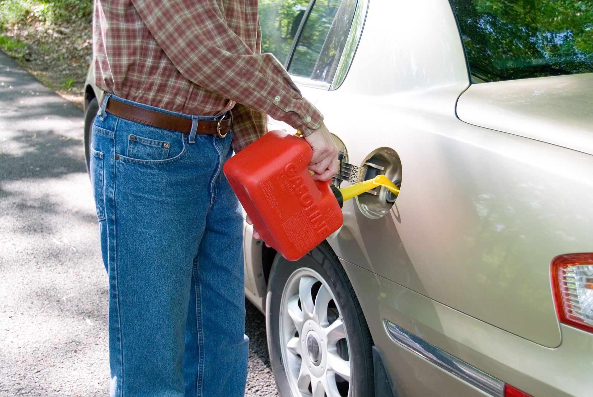 A person pours gasoline from a red plastic container into the fuel tank of a light-colored sedan parked on a roadside.