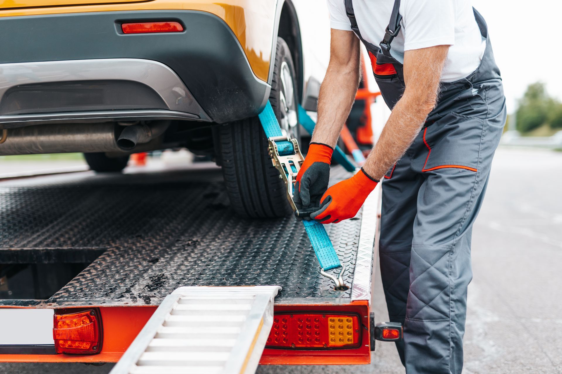 A person wearing orange gloves and gray work clothes secures a car to a flatbed tow truck using a blue ratchet strap.