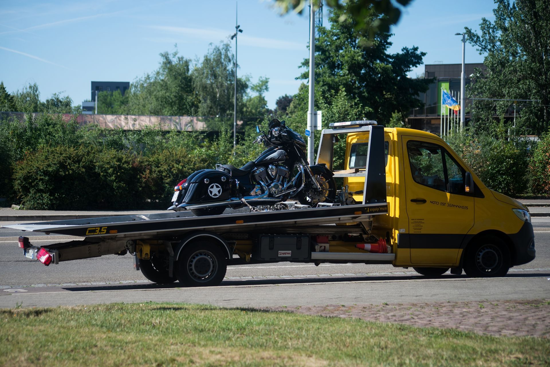 A yellow flatbed tow truck transporting a black motorcycle on an asphalt road near greenery.