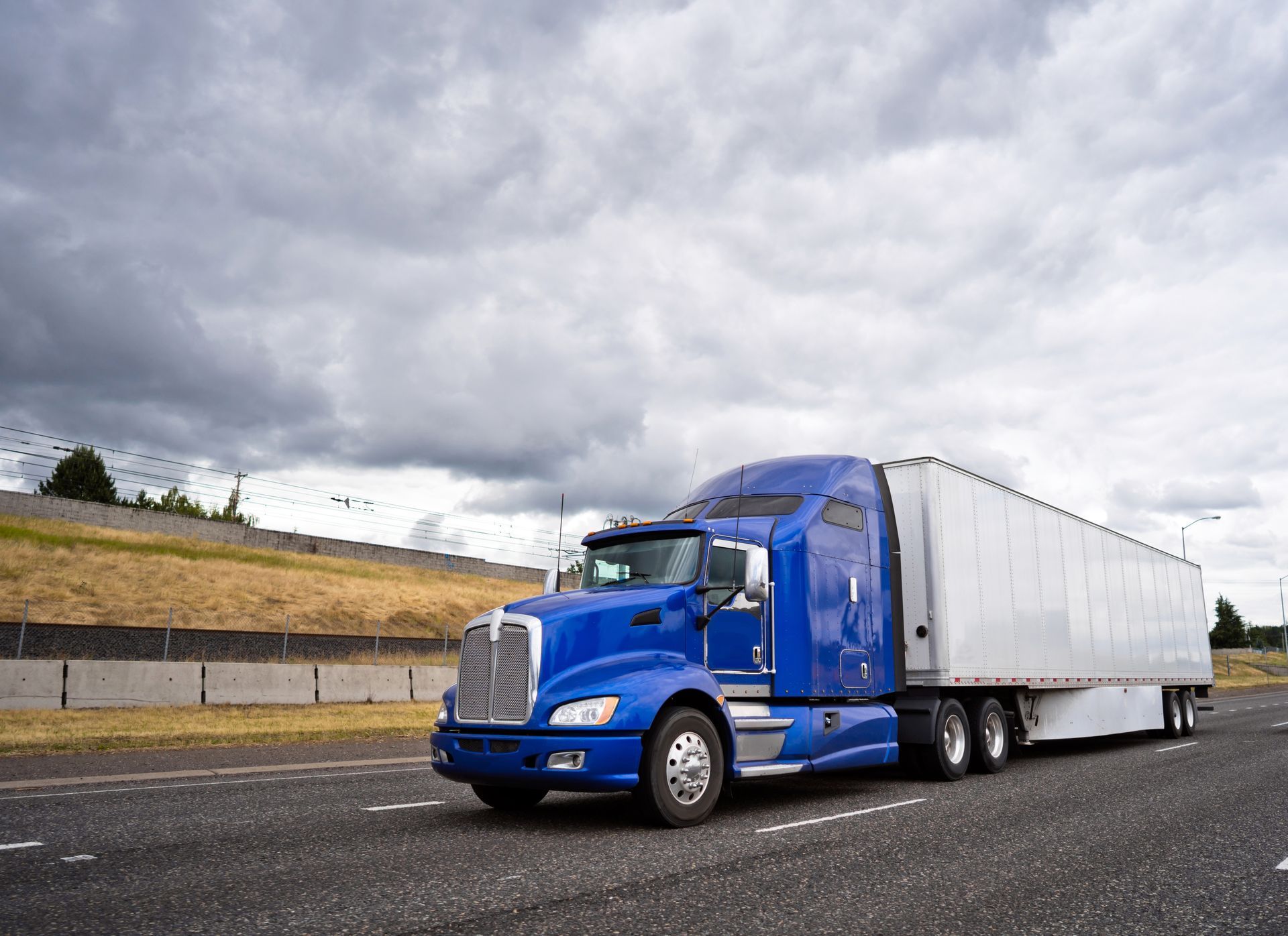 A blue semi-truck with a white trailer driving on an asphalt highway under a cloudy sky.