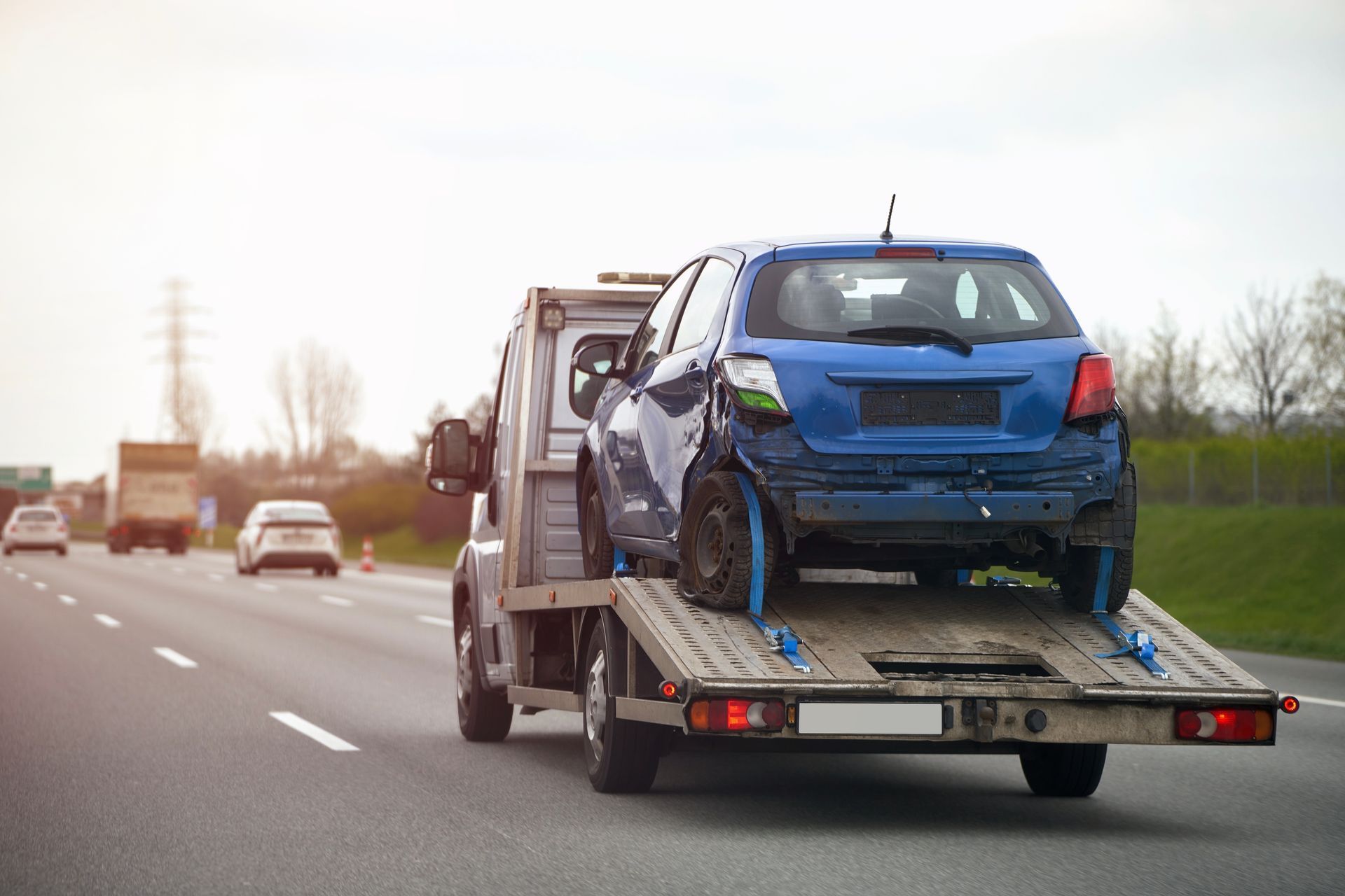 A damaged blue hatchback car being transported on a flatbed tow truck on a highway.