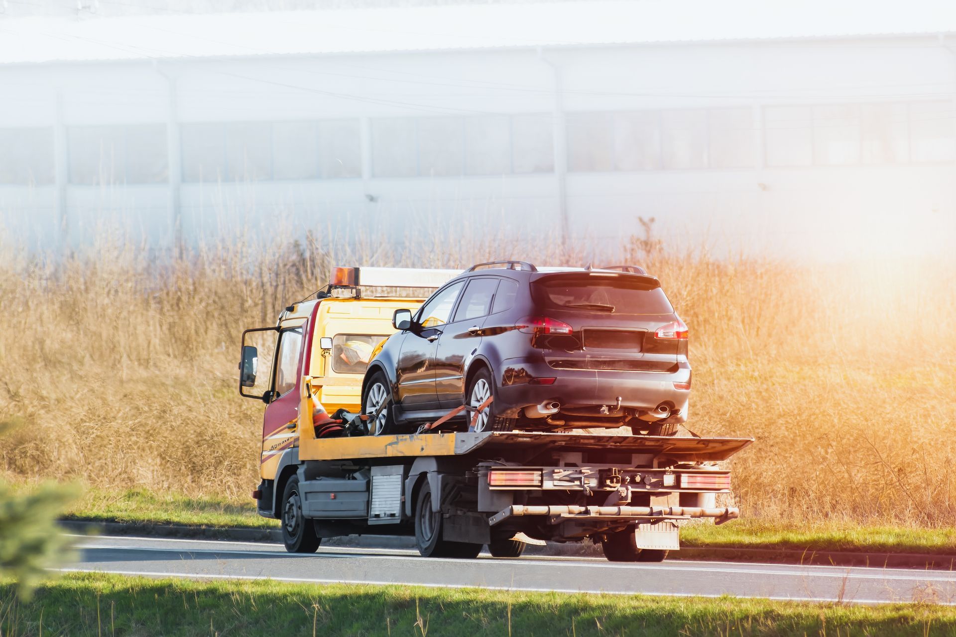 A dark car being transported on the back of a yellow tow truck along a road with tall, dry grass in the background.