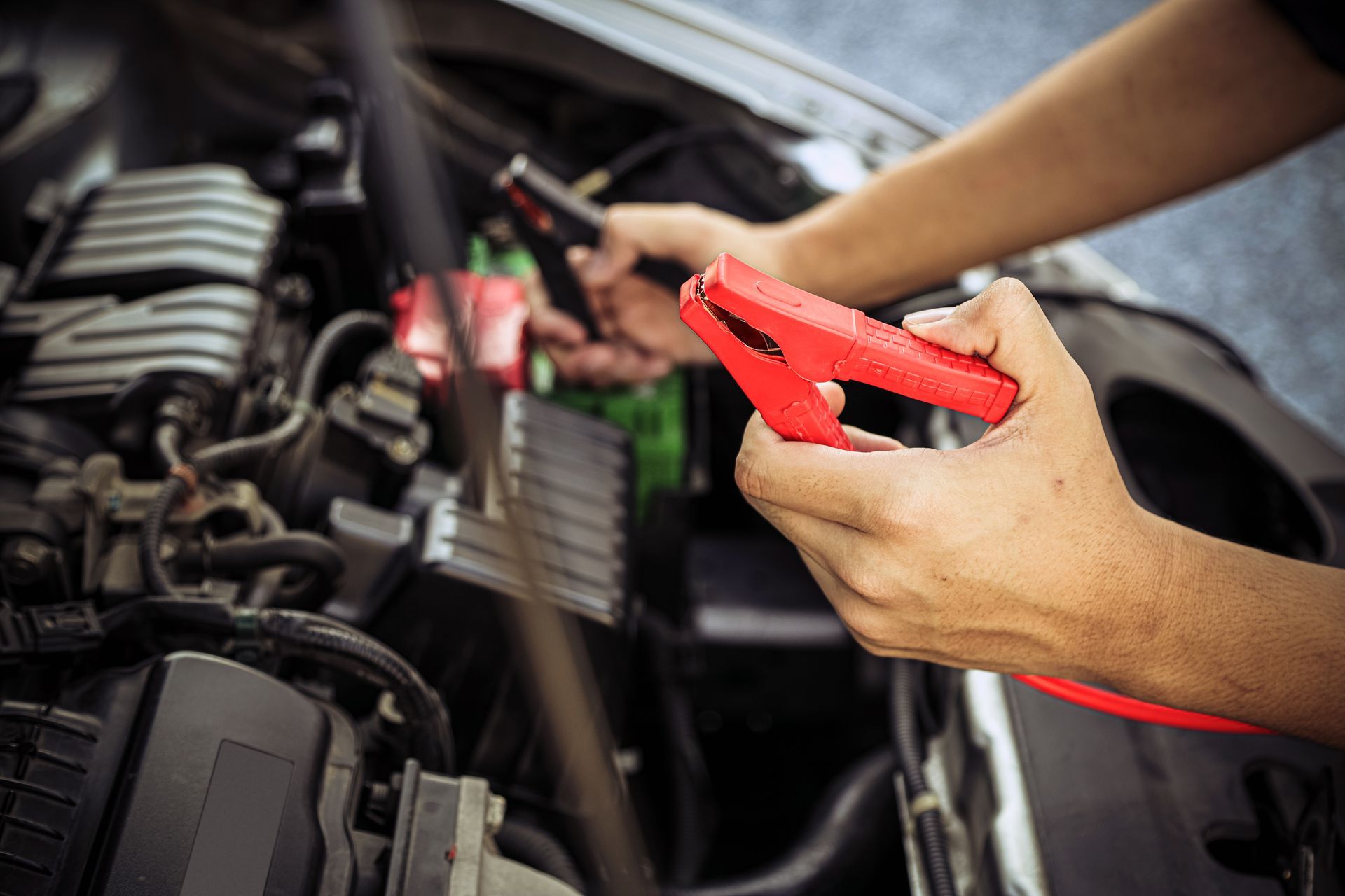 A pair of hands holds a red jumper cable clamp near an open car engine bay.