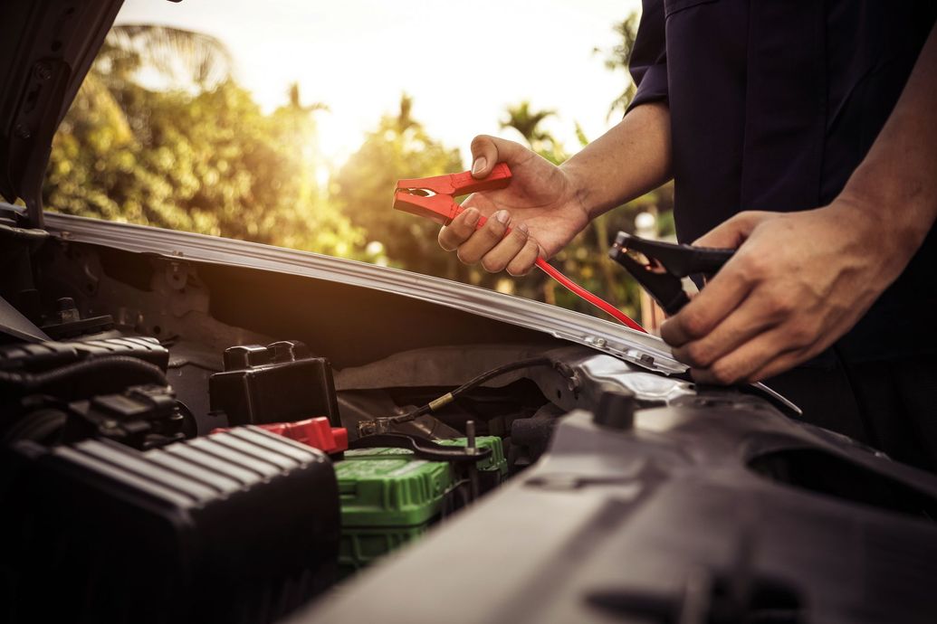 A person holding red and black jumper cable clamps over an open car engine, preparing to jump-start a vehicle battery.