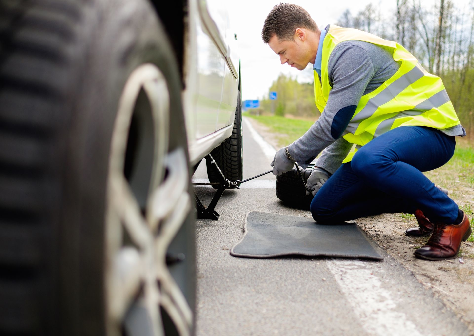 A person in a high-visibility vest kneels by a car on a road, using a jack to lift the vehicle for a tire change.